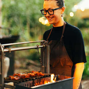 A person in an apron grills skewered food over an outdoor fire, smiling pleasantly.