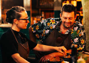 Two people in aprons prepare food at a lively outdoor food stall.