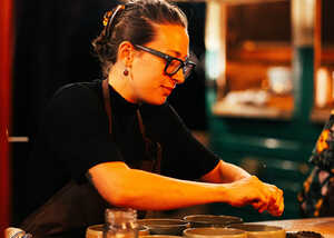 A woman in glasses prepares food at a table in a dimly lit setting.